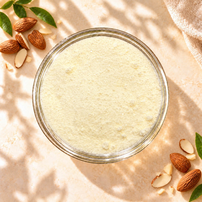 Top view of Almond Flavour Powder in a clear glass bowl, showing its fine, pale cream powder texture with whole almonds and almond slices on a warm beige background.