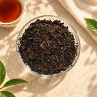 Loose Assam black tea leaves in a clear glass bowl, showing their dark brown colour and dried, twisted leaf texture, with brewed black tea beside them.