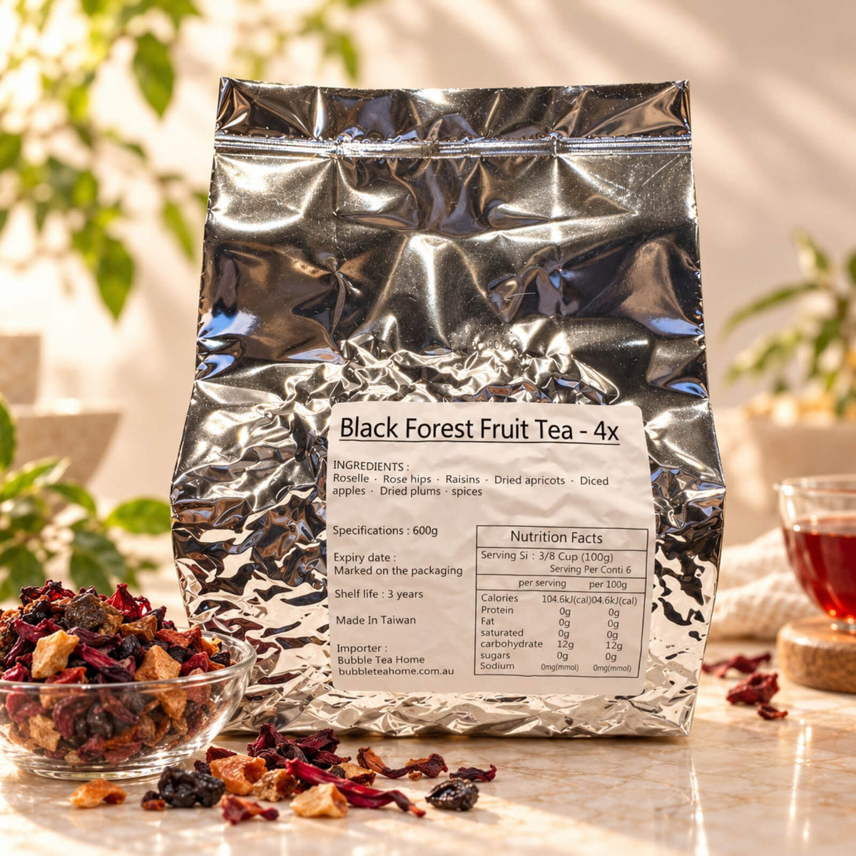 600g package of Black Forest Fruit Tea  in a silver foil bag with a product label, displayed alongside a bowl of dried fruit tea blend and brewed red fruit tea.