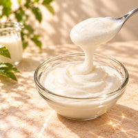 Thick and creamy cheese milk foam being lifted with a spoon above a clear glass bowl, showing its smooth, fluffy texture on a warm beige background.