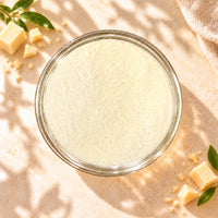 Top view of Cheese Milk Foam Powder in a clear glass bowl, showing its fine, pale cream texture with small cheese pieces on a warm beige background.