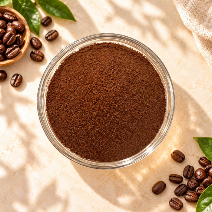 Top view of Coffee Powder in a clear glass bowl, showing its rich brown colour and fine texture, surrounded by roasted coffee beans on a warm beige background.