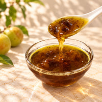 Golden crispy plum syrup pouring from a ceramic spoon into a glass bowl, showing whole preserved plums and thick golden syrup with fruit pieces.