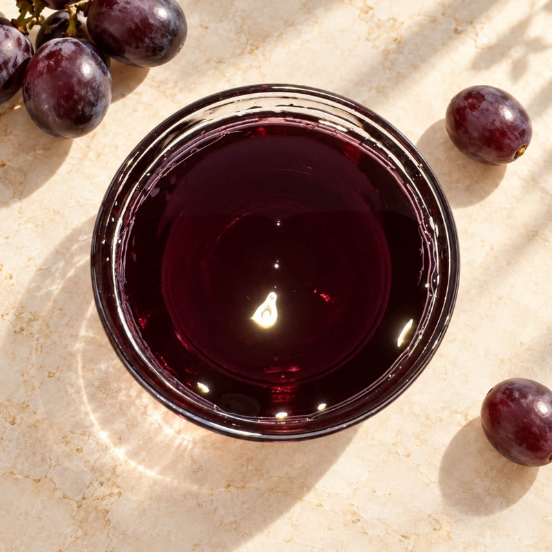Dark burgundy grape flavoured syrup in a glass bowl, showing its rich colour and smooth, glossy texture, surrounded by red grapes.