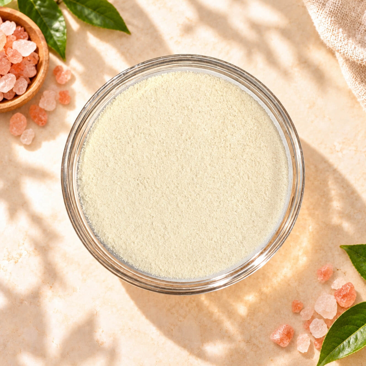 Top view of Himalayan Pink Salt Milk Foam Powder in a clear glass bowl, showing its fine creamy texture, surrounded by pink salt crystals on a warm beige background.