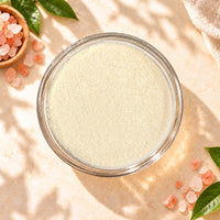 Top view of Himalayan Pink Salt Milk Foam Powder in a clear glass bowl, showing its fine creamy texture, surrounded by pink salt crystals on a warm beige background.