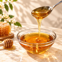 Golden amber honey flavoured syrup pouring from a spoon into a glass bowl, showing its thick, smooth and glossy consistency.