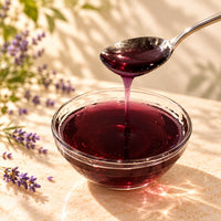 Deep purple-red lavender flavoured syrup pouring from a spoon into a glass bowl, highlighting its rich colour and thick, glossy consistency.