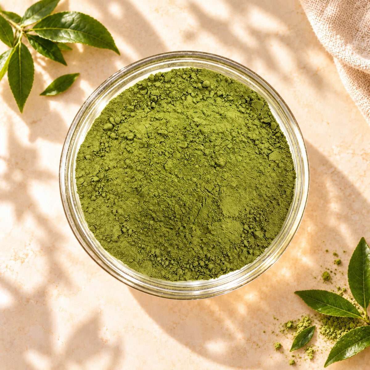 Top view of green Matcha Powder in a clear glass bowl, showing its fine texture, surrounded by tea leaves on a warm beige background.