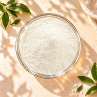 Top view of Non-Dairy Creamer Powder in a clear glass bowl, showing its fine, white powder texture on a warm beige surface with soft natural sunlight and leafy shadows.