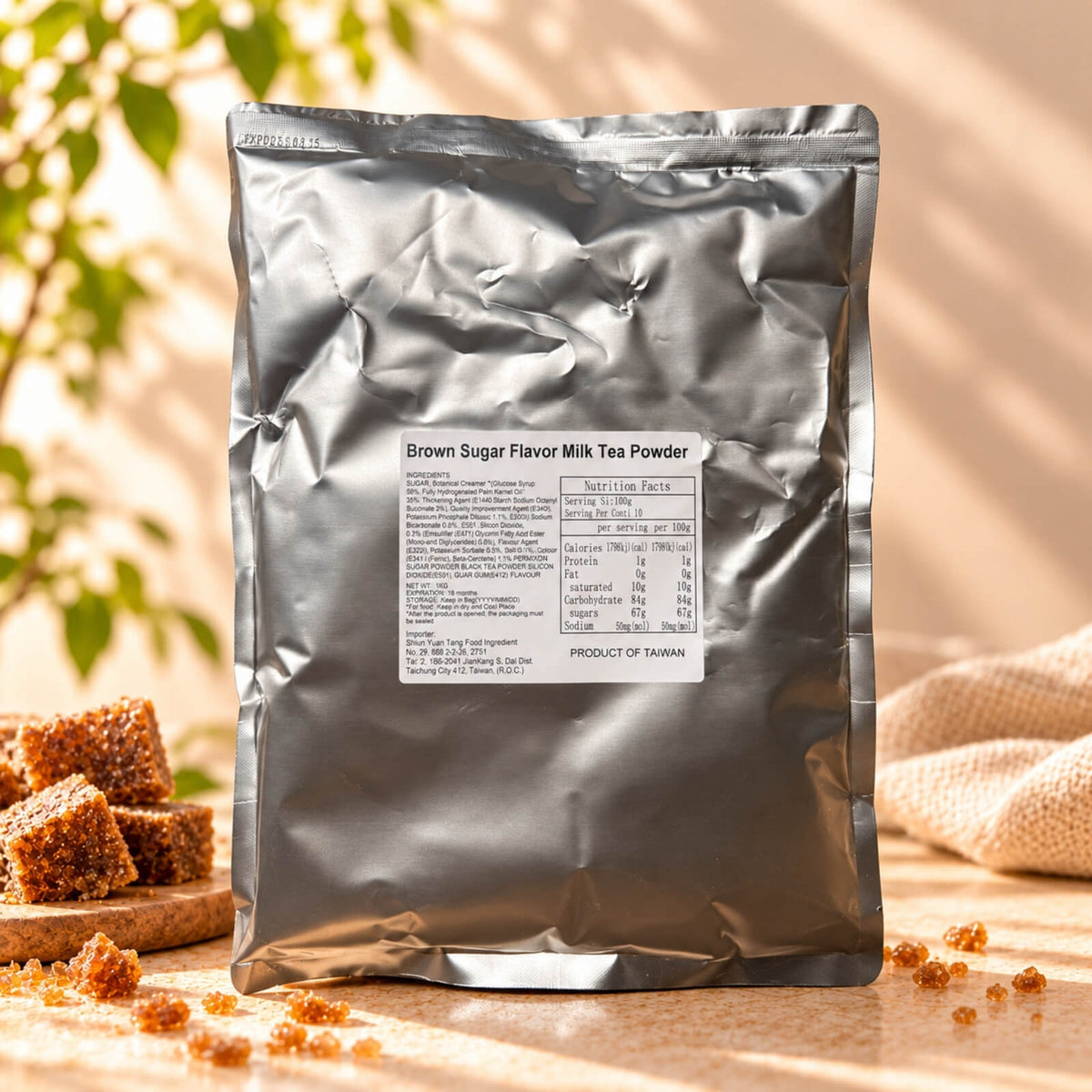 Okinawa Brown Sugar Milk Tea Flavour Powder in a silver foil package with a product label, displayed alongside brown sugar cubes under warm natural sunlight.