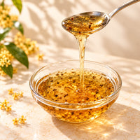 Golden osmanthus flavoured syrup pouring from a spoon into a glass bowl, showing its thick, glossy texture and delicate osmanthus flowers.