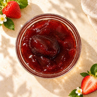 Strawberry jam in a glass bowl, showing its deep red colour, thick glossy consistency and strawberry fruit pieces, displayed alongside fresh strawberries.