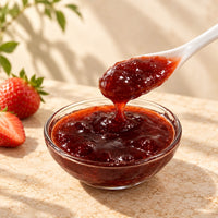 Thick strawberry jam dripping from a white ceramic spoon into a glass bowl, showing its rich red colour, glossy texture and visible strawberry pieces.