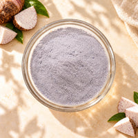 Top view of fine, light purple taro powder in a clear glass bowl, surrounded by fresh taro roots and slices on a warm beige surface.